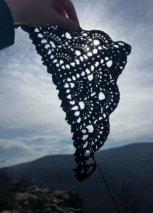 Black crochet bandana held up against a cloudy sky and landscape background.