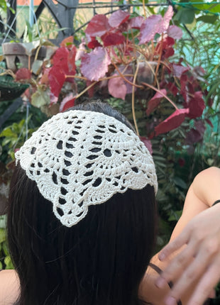 Back view of a white crochet bandana worn in dark hair near garden foliage.